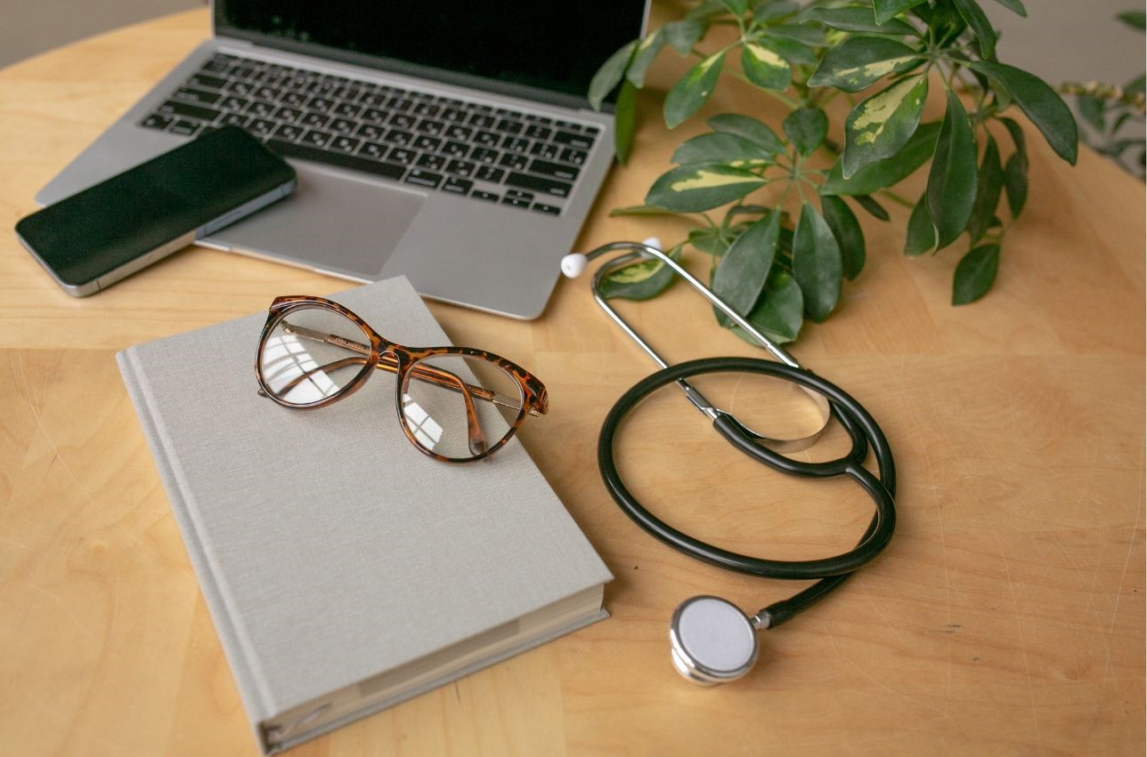 Imagen de un ordenador, un teléfono móvil, un libro, unas gafas, un estetoscopio y una planta sobre una mesa de trabajo